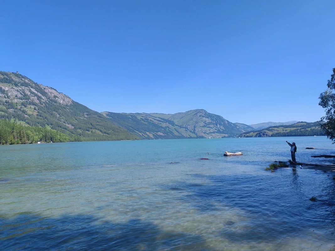 The blue surface of Kanas Lake in the Chinese Altai in summer, with forested mountains rising on both sides and a single person standing on the rocky shore pointing toward the opposite ridge