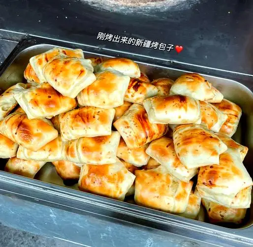 A stainless steel tray piled with Xinjiang baked samsa, the pastry golden-brown and pinched into the traditional rectangular fold, with Chinese characters and a red heart drawn on the steam of the glass above